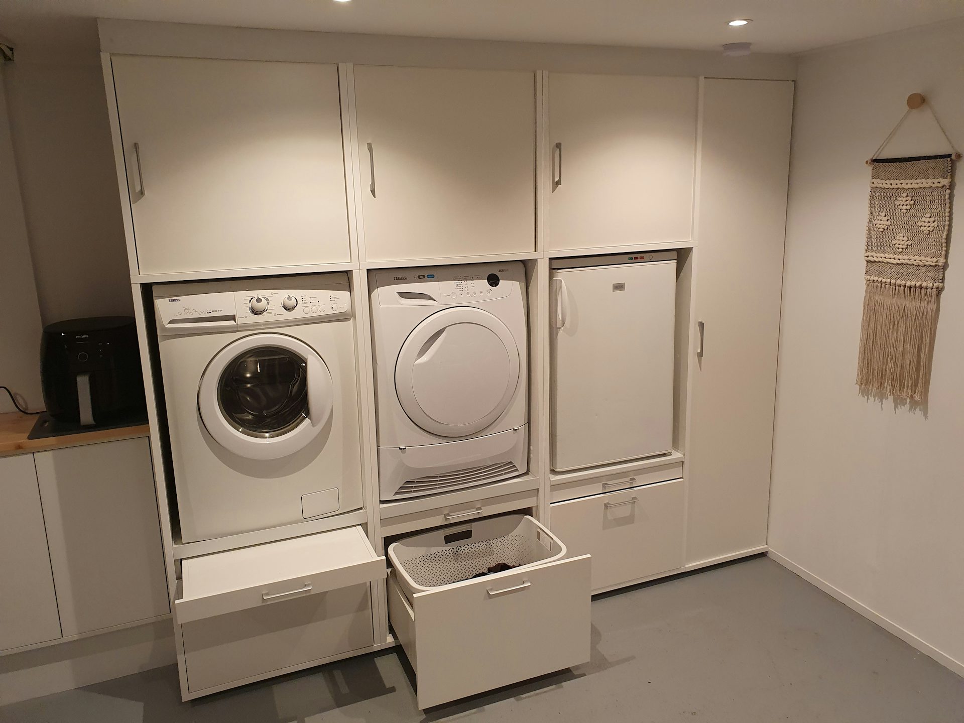Voorbeeld witte kastenwand opstelling in washok met wasmachine, droger, koelkast, ladekast, opbergruimte en uittrekplateau voor de wasmand